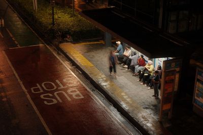 A dimly lit bus stop at night with diverse passengers waiting under shelter.